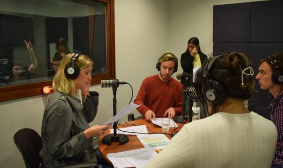 Cinco estudiantes en un estudio de radio, con auriculares en torno a una mesa. Se ve una ventana y del otro lado el operador levanta la mano para indicar algo. 