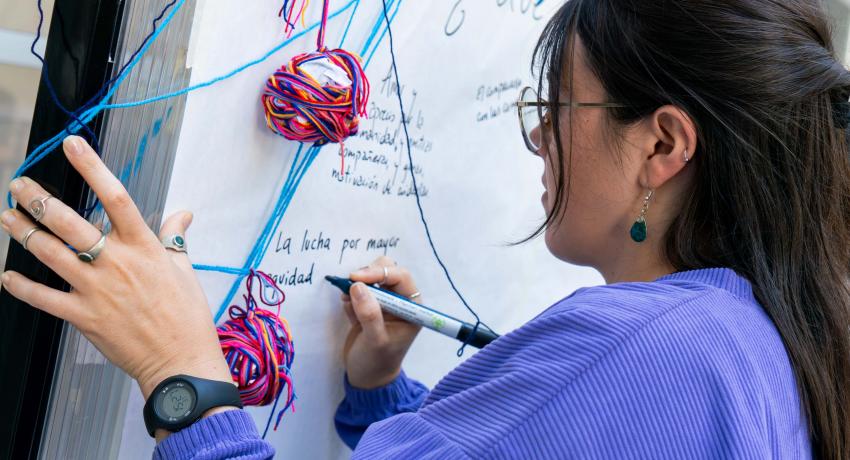 Fotografía. Encuentro de Mujeres del ASA. Una de las participantes escribe en un papelógrafo. Plano medio, de perfil, de pelo largo oscuro y lentes, lleva buzo azul. Se atraviesan hilos de lana celeste, azul y del papelógrafo cuelgan dos pombones de hilos de disntintos colores (rojos, rosados, azules).