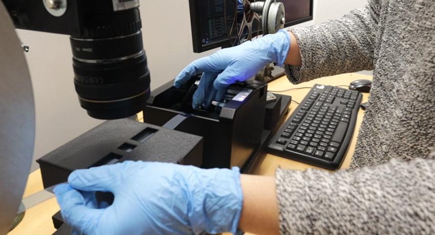 Foto: digitalización de película en scaner. Una persona con guantes de latex celeste sostiene la cinta en el artefato por los extremos. 