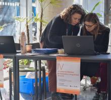Foto. Mesa de acreditaciones. Las docentes Victoria Cuadrado y Romina Escobar trabajan en  una computadora. Pegado desde el borde de la mesa hay dos afiches que dicen en letras color naranja sobre fondo blanco: "IV Jornadas Académicas de Extensión e Integralidad de la FIC"