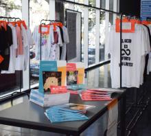 Foto de la presentación del libro de la UDEA. Sobre una mesa varios libros apilados. Detrás ven dos filas de remeras negras y blancas con inscripciones colgadas en percheros.