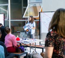 Foto. Encuentro de Mujeres del ASA. De espaldas cuatro participantes del encuentro atienden a una joven que expone su trabajo durante el taller de fanzine. 